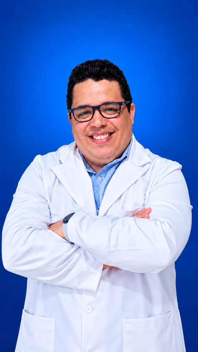 Professor Thiago Santos de jaleco branco, braços cruzados, em fundo azul